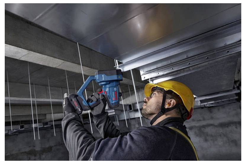 Ein Arbeiter mit einem gelben Schutzhelm verwendet ein Elektrowerkzeug, um Metallkanäle an einer Decke zu befestigen und damit Bau- oder Installationsarbeiten zu veranschaulichen.