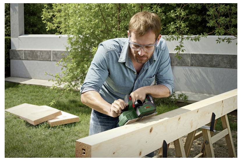 Ein Mann mit Schutzbrille bearbeitet einen Holzbalken mit einem Elektrowerkzeug im Freien, wobei sich im Hintergrund weiteres Holz und Grünpflanzen befinden.