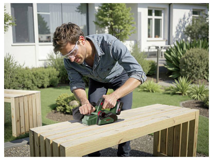 Eine Person mit Sicherheitsbrille benutzt ein Elektrowerkzeug, um eine Holzbank im Freien zu hobeln, umgeben von Pflanzen und einem Gebäude im Hintergrund.