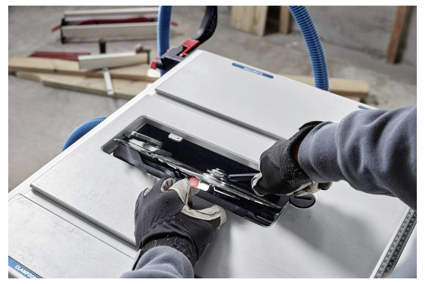 Eine Person mit Handschuhen justiert die Klinge einer Kreissäge, an der ein blauer Schlauch angeschlossen ist. Holzstücke und ein Messwerkzeug befinden sich im Hintergrund.