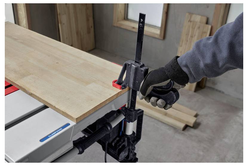Eine Person mit Handschuhen befestigt mit einer Klemme ein Holzbrett auf einer Werkbank in einer Werkstattumgebung.
