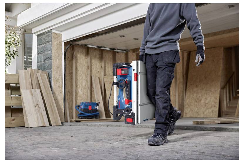 Eine Person in Arbeitskleidung trägt einen blauen und roten Bosch-Werkzeugkoffer einen Einfahrtsweg entlang vor einer offenen Garage, in der Holzbretter liegen.