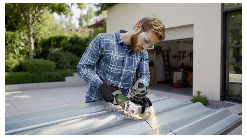 Eine Person mit Schutzbrille und kariertem Hemd verwendet einen Elektrowinkelschleifer, um Metallbleche im Freien in der Nähe einer Garage zu schneiden.