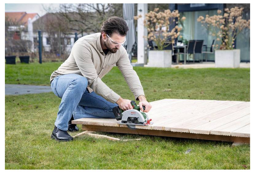 Eine Person mit Schutzbrille verwendet eine Kreissäge, um Holzbretter auf einem Rasengrundstück zu schneiden, mit einem modernen Haus im Hintergrund.