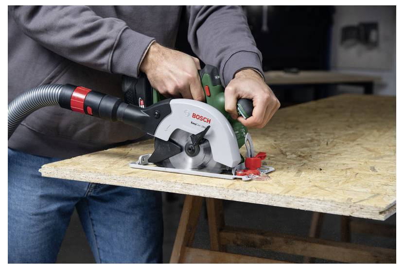 Eine Person verwendet eine Bosch-Kreissäge, um eine Holzplatte auf einer Werkbank zu schneiden. Der angeschlossene Saugschlauch sammelt Staub.