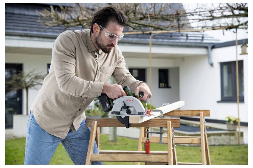 Mann, der eine Kreissäge verwendet, um Holz auf einem Sägebock außerhalb eines modernen Hauses zu schneiden.