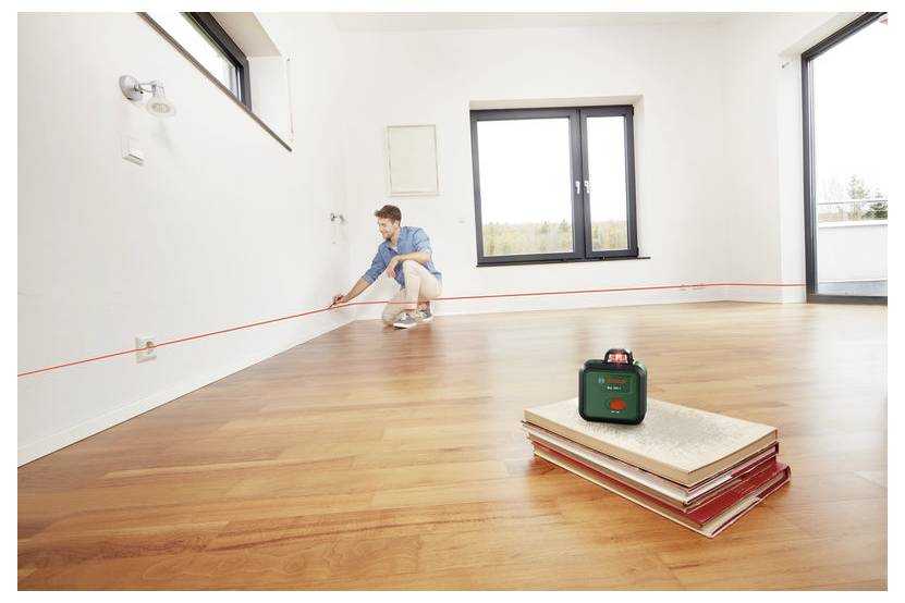 Eine Person in einem Raum verwendet eine Wasserwaage mit Laser, um eine gerade Linie an der Wand auszurichten. Ein Stapel Bücher im Vordergrund stützt das Lasergerät.