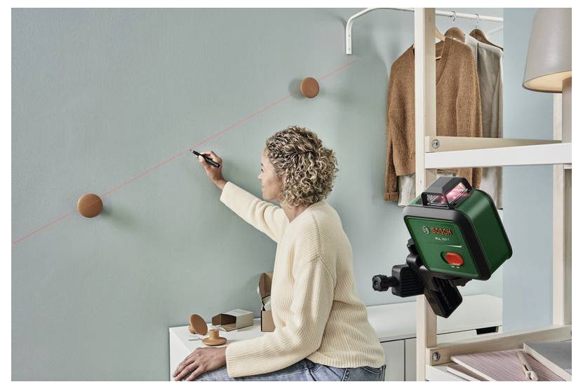 Eine Person verwendet eine Laserwasserwaage, um eine gerade Linie an einer Wand in einem Raum zu ziehen, wobei beige Kleidungsstücke an einem Ständer in der Nähe hängen.