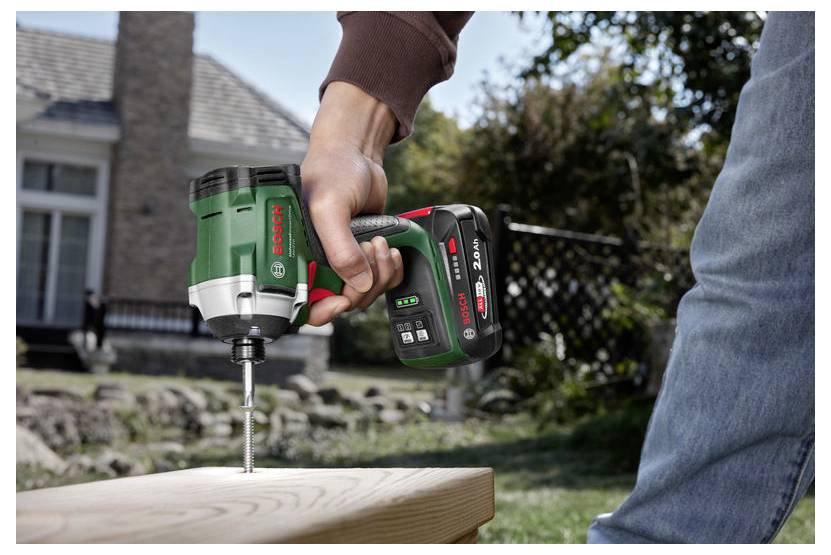 Eine Person verwendet einen kabellosen elektrischen Schraubendreher, um eine Schraube in ein Holzbrett im Freien zu bohren, wobei ein Haus im Hintergrund zu sehen ist.