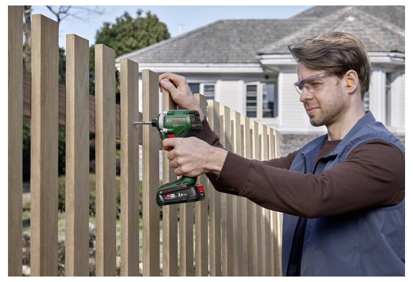 Ein Mann mit Schutzbrille verwendet eine kabellose Bohrmaschine, um Holzbretter an einem Zaun in einem Wohnhinterhof zu befestigen.