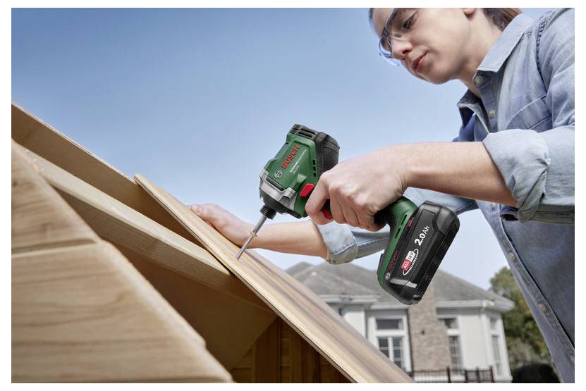 Eine Person verwendet einen Bosch-Akku-Bohrer, um Holzplanken auf einem Dach zu befestigen, mit einem Haus im Hintergrund und unter Verwendung einer Schutzbrille.