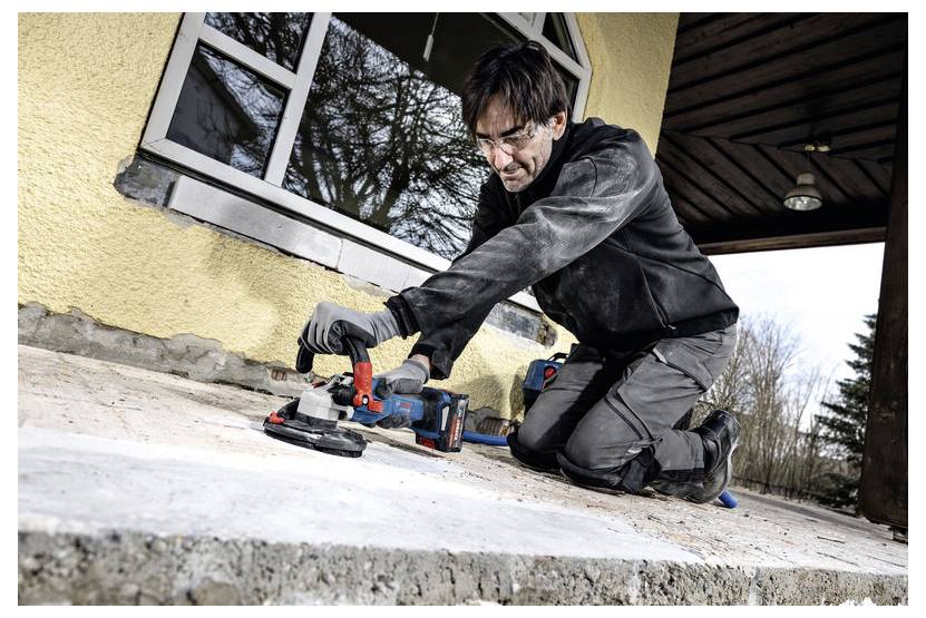 Eine Person in Schutzausrüstung kniet auf einer Betonoberfläche und verwendet ein Elektrowerkzeug zum Schleifen oder Polieren. Ein Haus und Bäume sind im Hintergrund zu sehen.