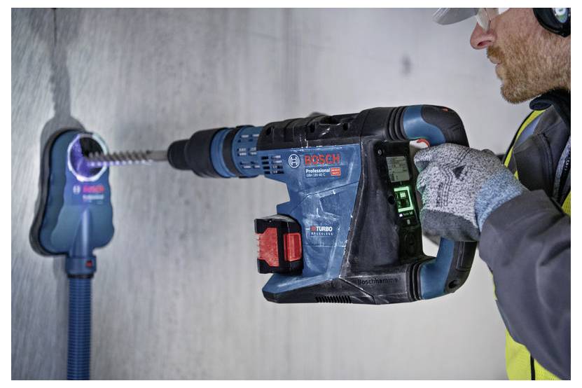Eine Person in Schutzausrüstung verwendet einen Bosch Professional Akku-Bohrer an einer Betonwand und demonstriert dessen Staubabsaugfunktion.