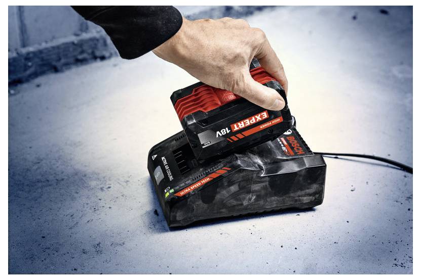 Eine Person legt einen roten Bosch 'Expert 18V' Akku auf eine schwarze Ladestation auf einem Betonboden. Die Ladestation verfügt über Anzeigen und ein Netzkabel.