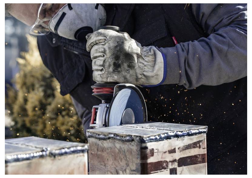 Eine Person trägt Schutzhandschuhe und Schutzbrille und verwendet einen Winkelschleifer, wobei Funken entstehen, während sie durch Metall schneidet.