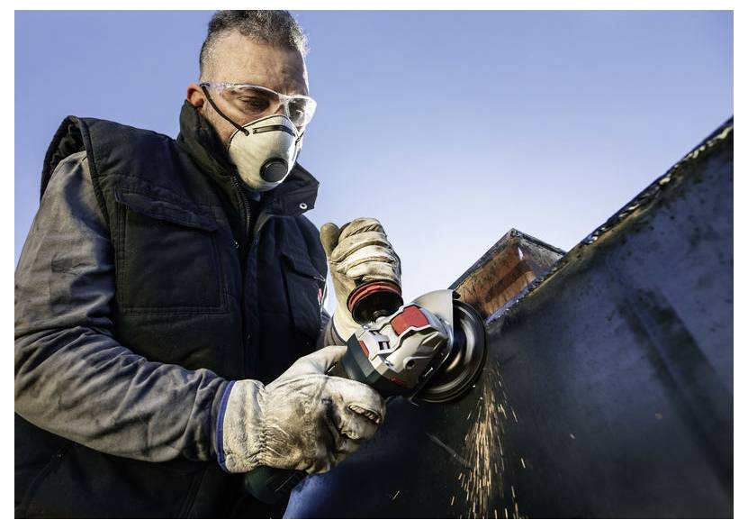 Eine Person mit Schutzbrille und Maske verwendet eine Winkelschleifer auf einer Metalloberfläche, wobei Funken sprühen, vor einem klaren blauen Himmel.