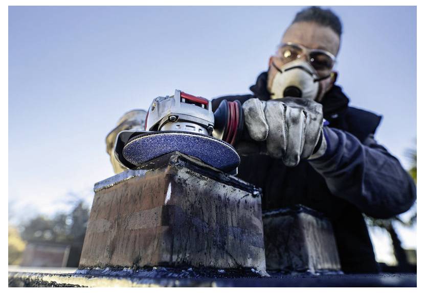 Eine Person mit Maske und Handschuhen verwendet ein Elektrowerkzeug zum Schleifen von Metall im Freien, vor einem blauen Himmel als Hintergrund.