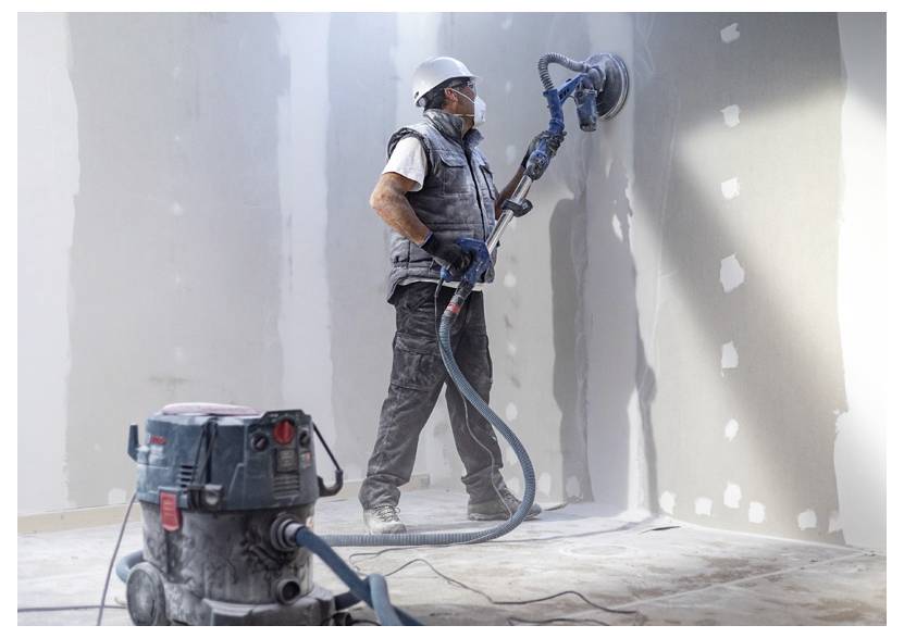 Ein Bauarbeiter mit Schutzausrüstung schleift eine Trockenbauplatten in einem Raum, der renoviert wird, und verwendet dabei einen Staubsauger, der an einen Schleifer angeschlossen ist.