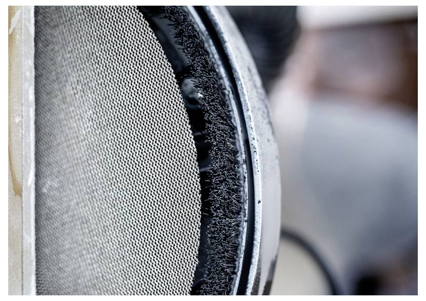 Nahaufnahme eines strukturierten Metallgitters mit einer kreisförmigen, dichten schwarzen Borstenreinigungsbürste am Rand, möglicherweise Teil eines Reinigungs- oder Filterungsmechanismus.