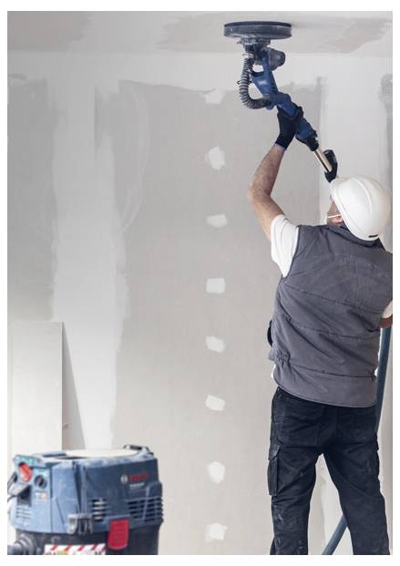 Arbeiter, der eine Decke mit einem Elektrowerkzeug schleift und eine Weste und einen Schutzhelm trägt, in einem Raum, der renoviert wird, mit unfertig verputzten Wänden.