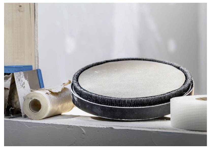 Ein Schleifwerkzeug mit einem weißen Oberflächenpad liegt auf einem Baustellenvorsprung, umgeben von einer blauen Kelle, Klebeband und einer Rolle Schleifpapier.