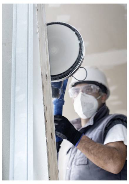 Ein Arbeiter mit Schutzhelm und Maske verwendet ein Elektrowerkzeug, um eine Trockenbauplatte in einer Baustelle zu schleifen.