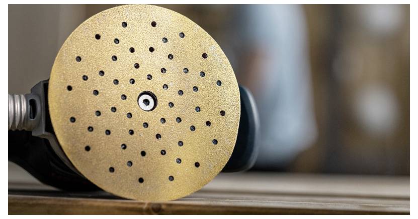Eine Nahaufnahme einer runden Schleifscheibe, die an einem Elektrowerkzeug befestigt ist und auf einer Holzoberfläche liegt, mit einem unscharfen Hintergrund.