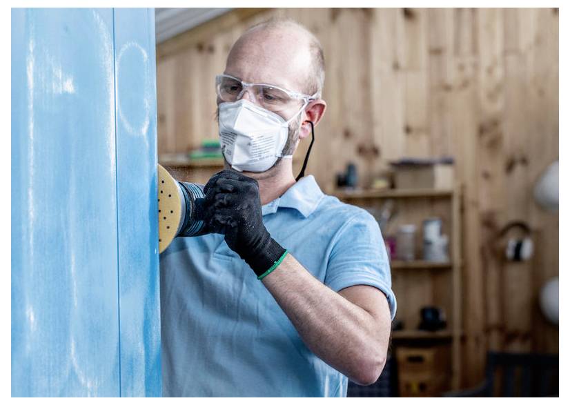 Eine Person trägt Schutzbrille, Maske und Handschuhe und verwendet einen Exzenterschleifer auf einer blauen Holzoberfläche, was auf Holzarbeit oder Möbelrestaurierung hindeutet.