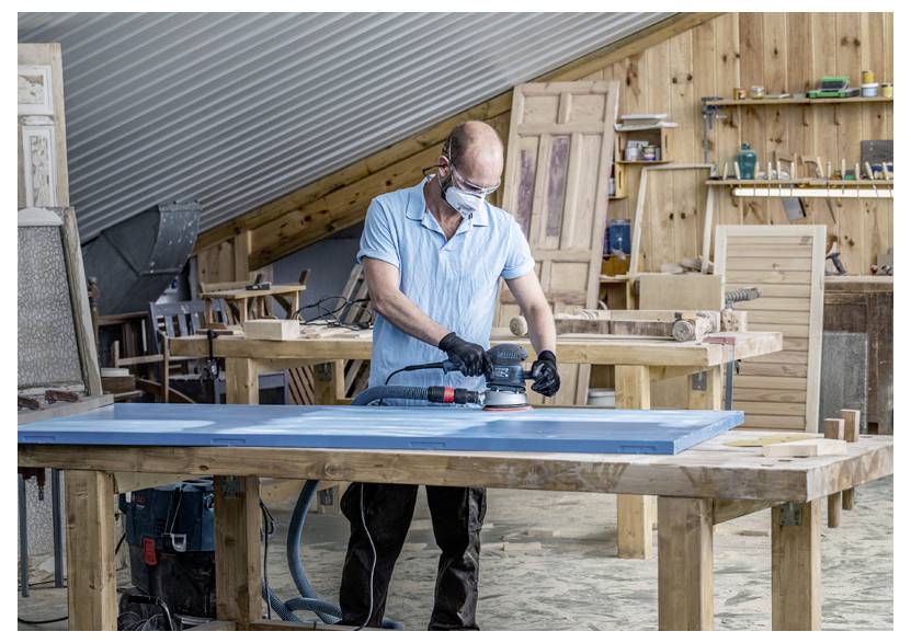 Eine Person mit Schutzbrille und Handschuhen schleift einen Holztisch in einer Werkstatt mit verschiedenen Werkzeugen und Holzplatten im Hintergrund.