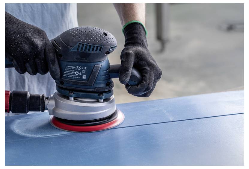 Eine Person mit Handschuhen benutzt einen kreisförmigen elektrischen Schleifer auf einer blauen Oberfläche und schleift diese gleichmäßig.