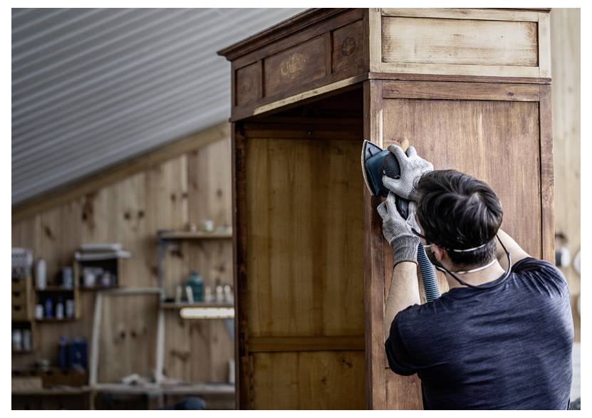 Eine Person mit Handschuhen und Atemschutzmaske schleift einen Holzschrank in einer Werkstatt, umgeben von Werkzeugen und Regalen.