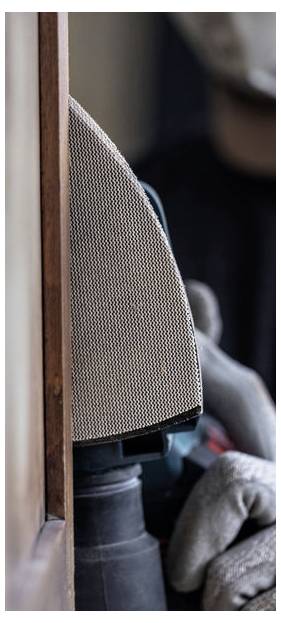 Eine Person mit Handschuhen verwendet einen Elektro-Schleifer auf einer Holzoberfläche und konzentriert sich dabei auf die Kante, was auf Holzarbeit oder Möbelrestaurierung hindeutet.