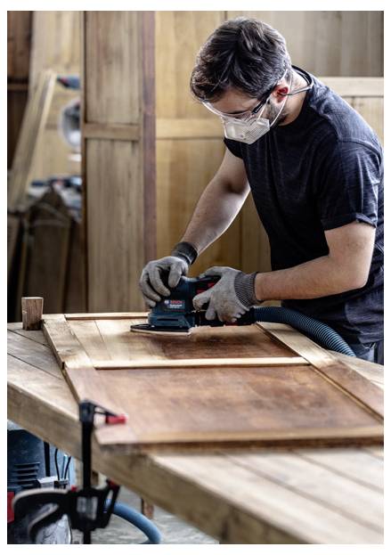 Eine Person mit Sicherheitsbrille und Maske schleift ein Holzbrett in einer Werkstatt.