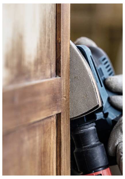 Eine Nahaufnahme einer Person mit Handschuhen, die einen Exzenterschleifer an der Kante eines Holzmöbelstücks verwendet und die Oberfläche glättet.