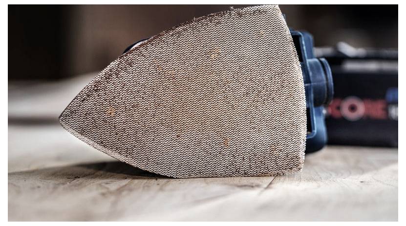 Ein Nahaufnahmebild einer Exzenterschleifmaschine auf einer Holzoberfläche, wobei der grobe, dreieckige Schleifpad hervorgehoben wird.