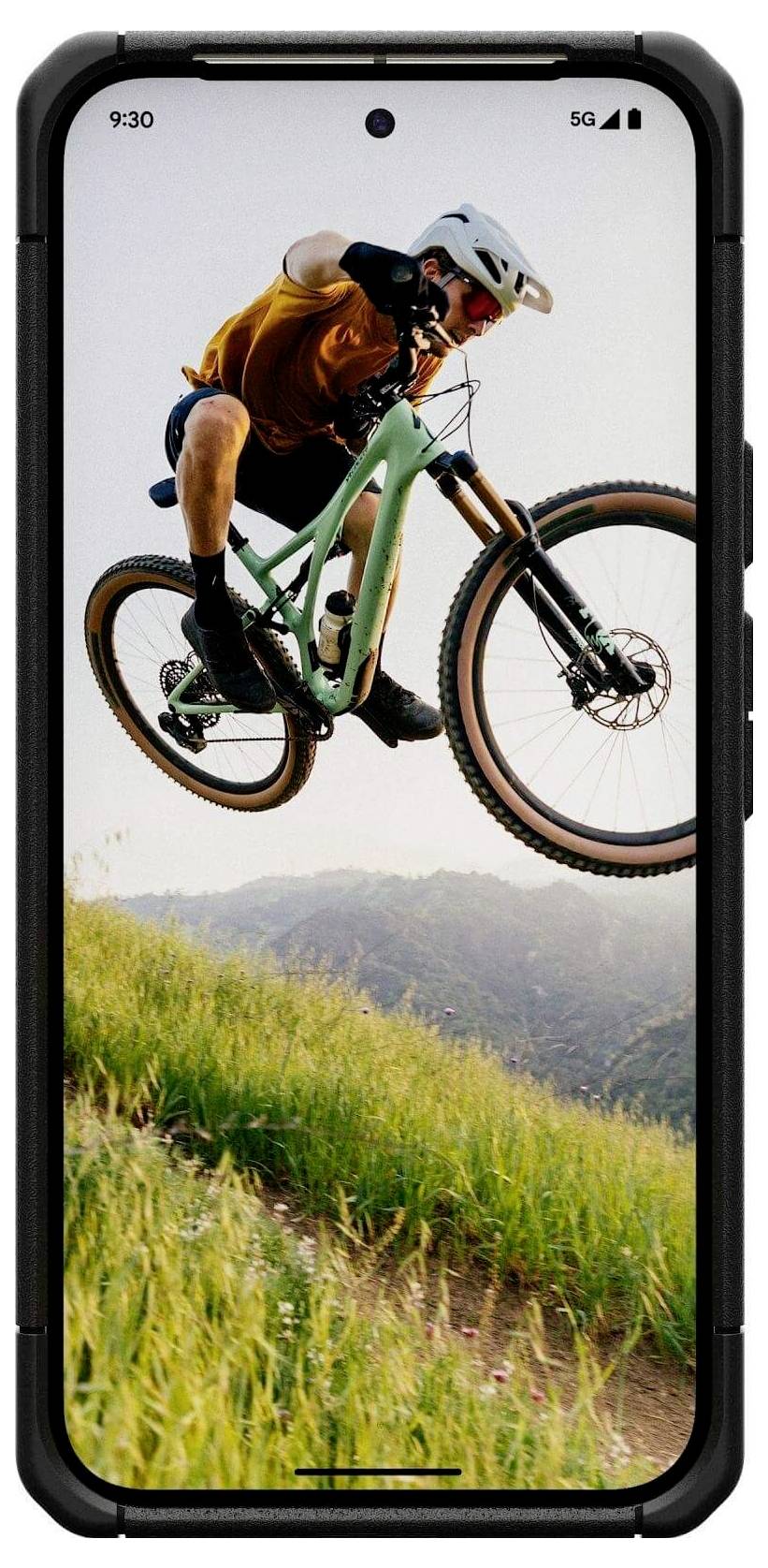 Ein Radfahrer in der Luft springt mit einem Mountainbike auf einem grasigen Hügel, aufgenommen auf einem Smartphone-Bildschirm, der die Kameraklarheit des Geräts zeigt.