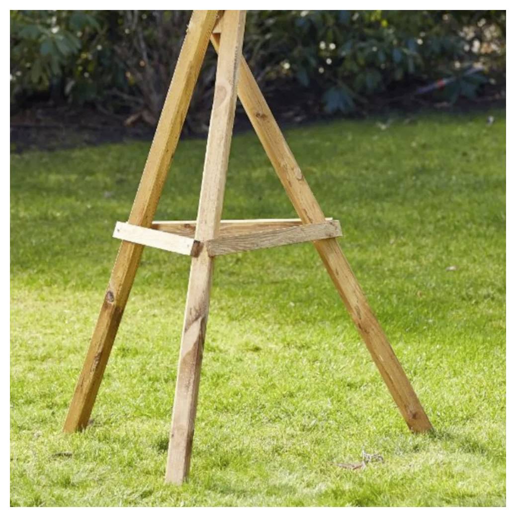 Hölzerne Staffelei, die auf grünem Gras in einer Gartenumgebung steht, mit belaubten Büschen im Hintergrund.