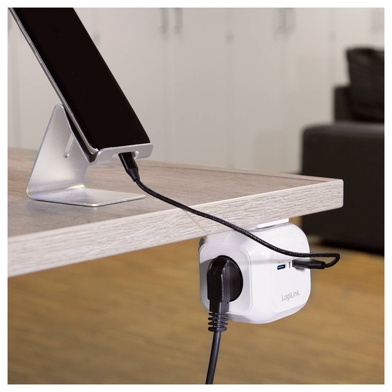 Ein Telefon auf einem Ständer wird über ein Kabel geladen, das mit einer Steckdose verbunden ist, die unter einem Holzschreibtisch montiert ist.