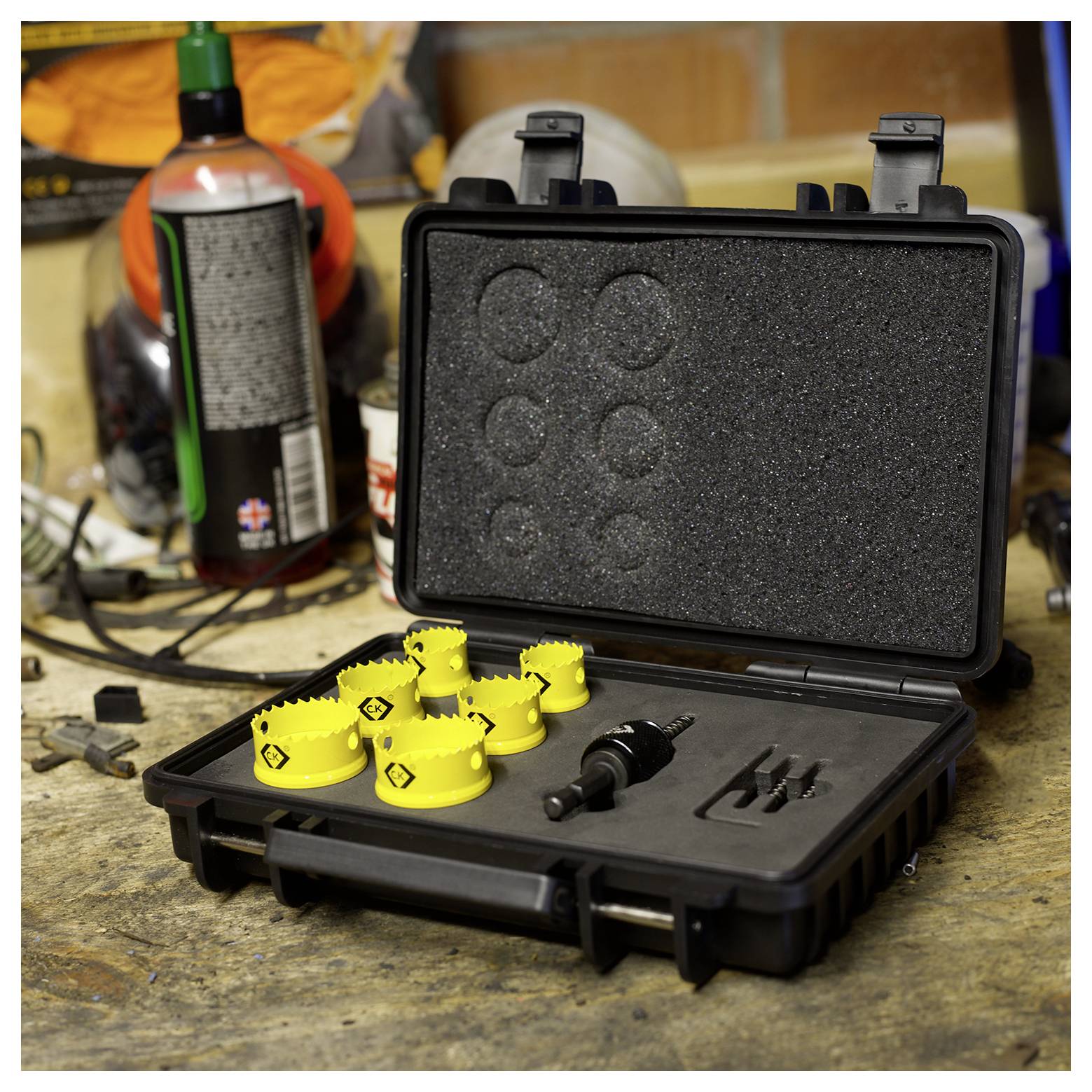 Eine schwarze Kiste mit offenem Deckel enthält einen gelben Lochsägesatz auf einer Werkbank, umgeben von Werkzeugen und Sprühflaschen. Ziegelwand im Hintergrund.