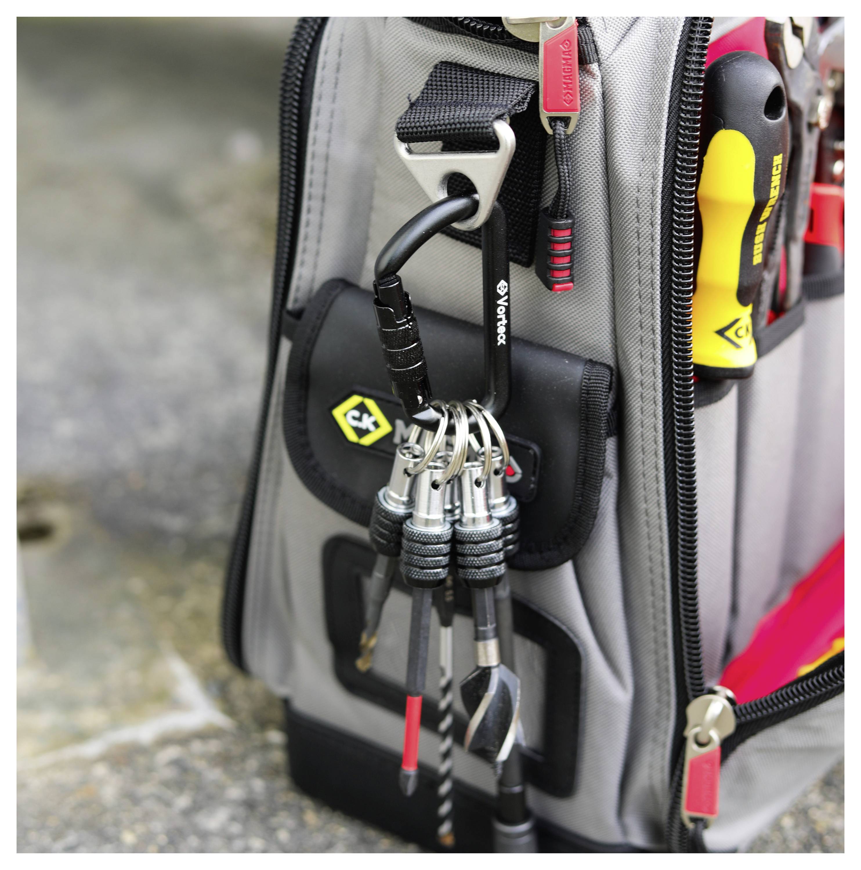 Eine Werkzeugtasche mit verschiedenen Handwerkzeugen und Zubehöranbringungen, einschließlich Schraubendreher und Bohrerspitzen, einsatzbereit auf einer Betonoberfläche.