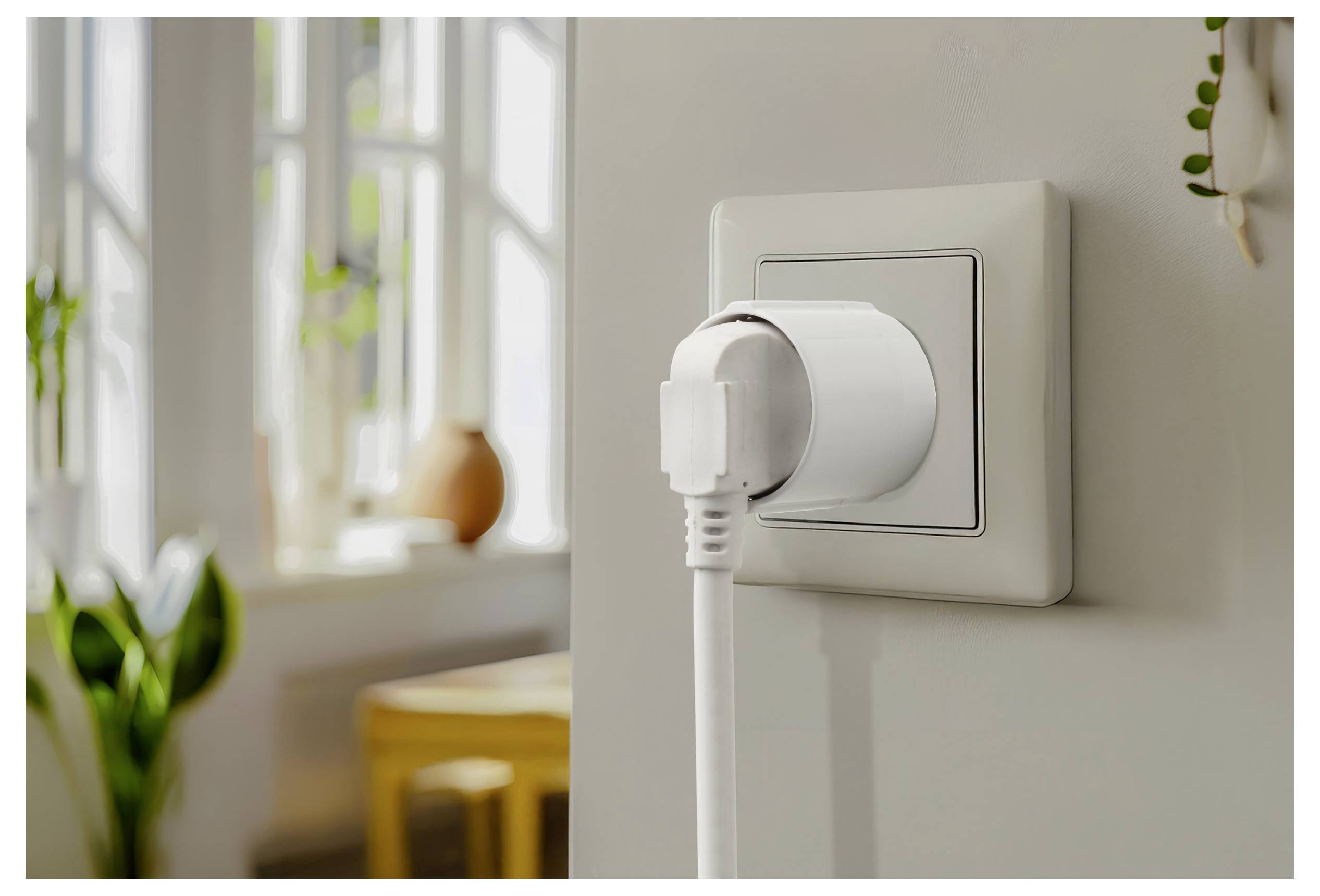 Ein weißer Netzstecker wird in eine Wandsteckdose in einem hellen Raum mit einem verschwommenen Hintergrund eingesteckt, der einen Tisch und ein Fenster zeigt.