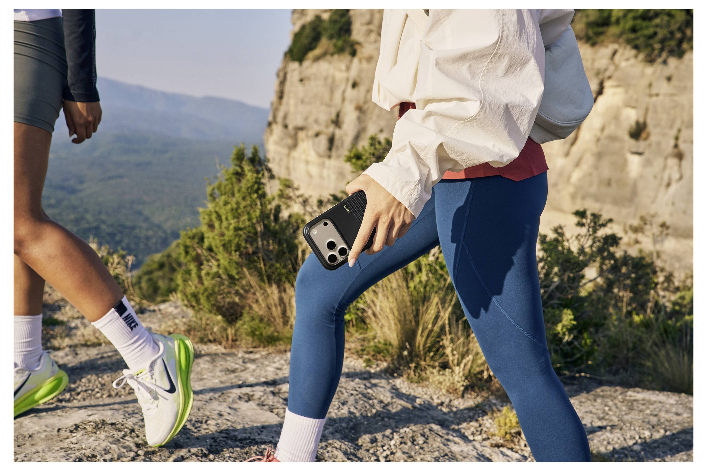 Zwei Personen wandern auf einem felsigen Pfad nahe einer Klippe, ein Smartphone in der Hand. Malerische Berge und Grünflächen im Hintergrund.
