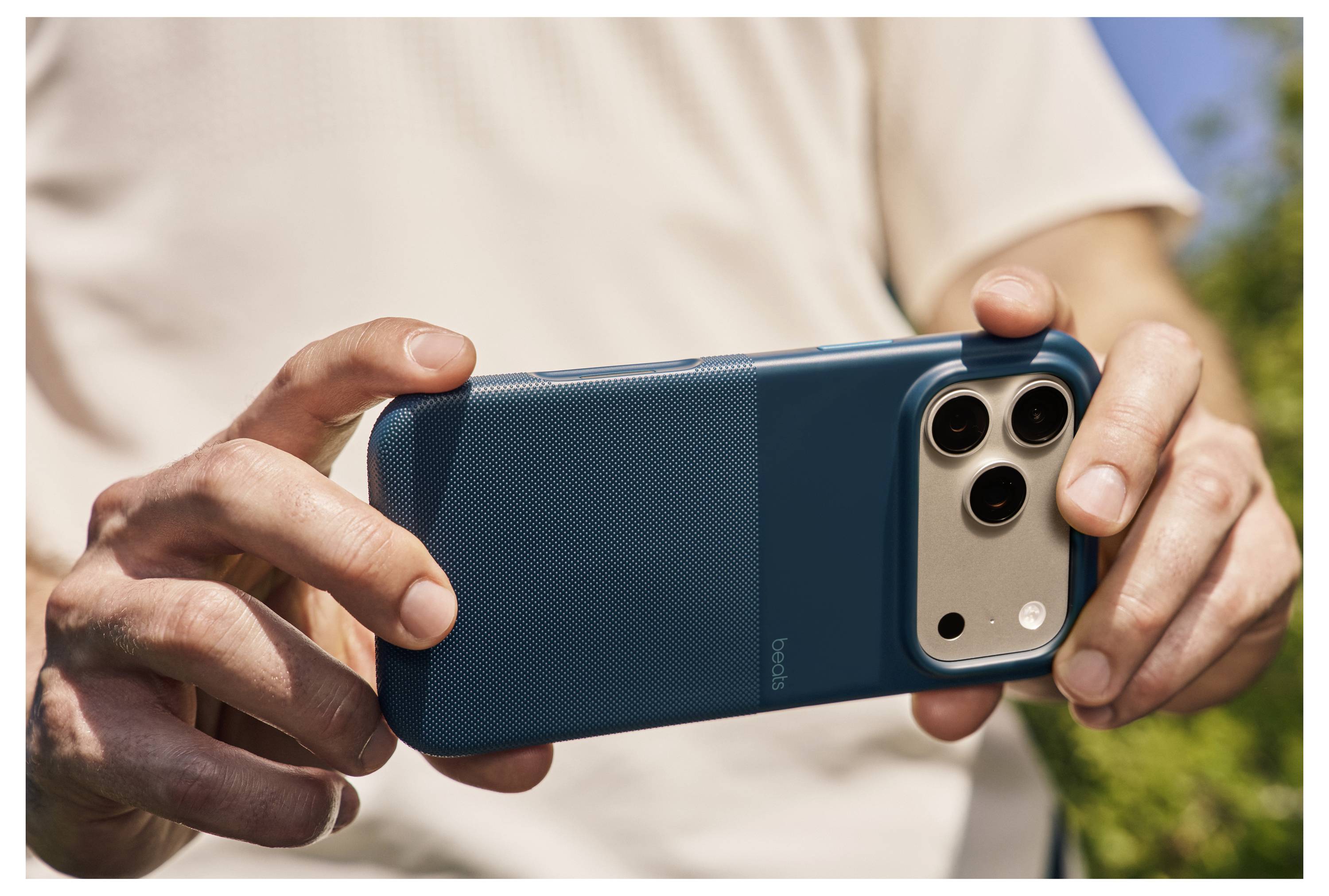 Eine Person hält ein Smartphone horizontal mit einer türkisfarbenen Hülle und richtet die Kamera aus. Outdoor-Umgebung mit unscharfem grünem Hintergrund.