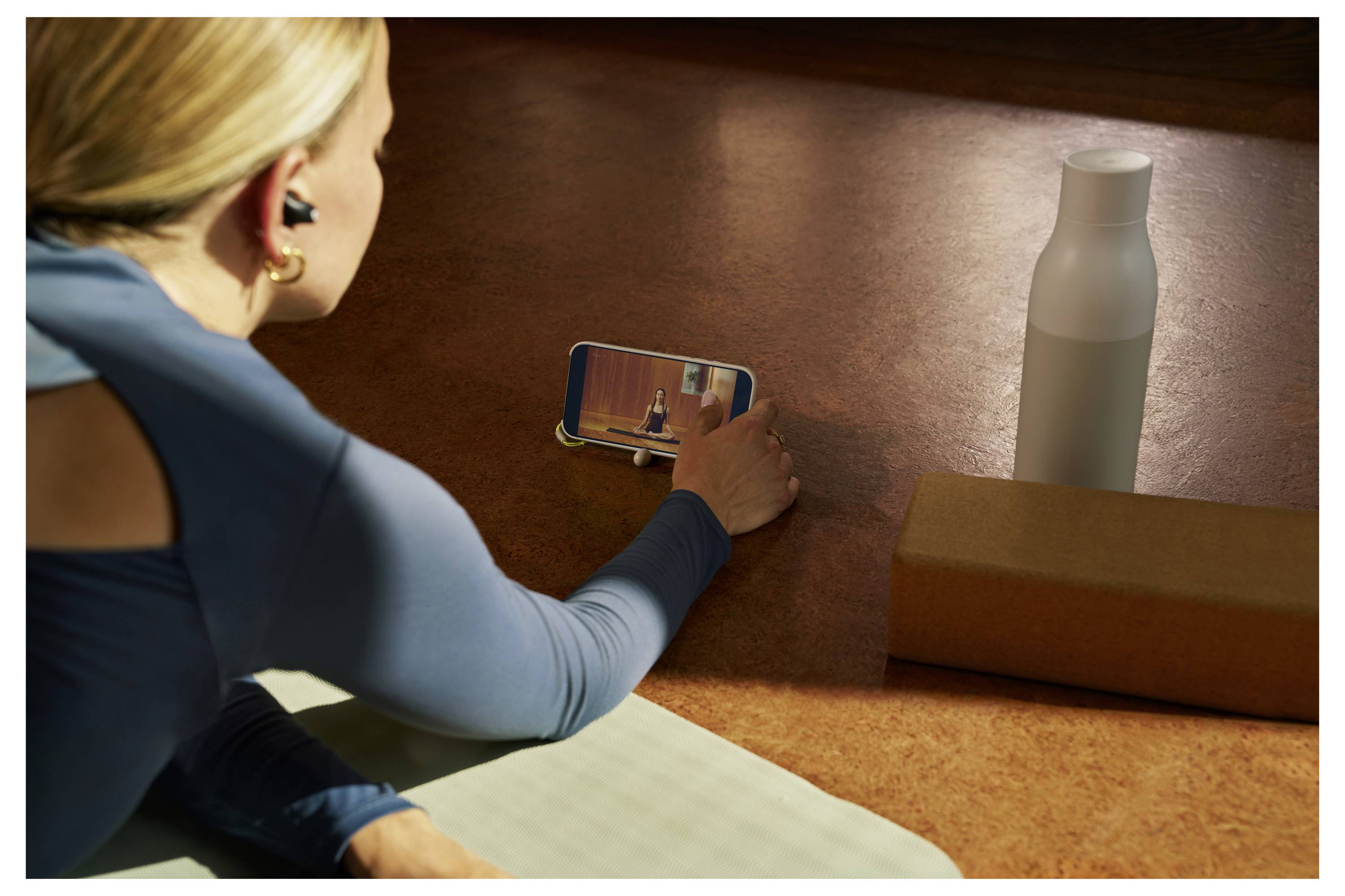 Eine Person, die einen Online-Yoga-Kurs zu Hause macht, liegt auf einer Yogamatte und schaut auf einen Smartphone-Bildschirm. Eine Wasserflasche und ein Yogablock befinden sich in der Nähe.