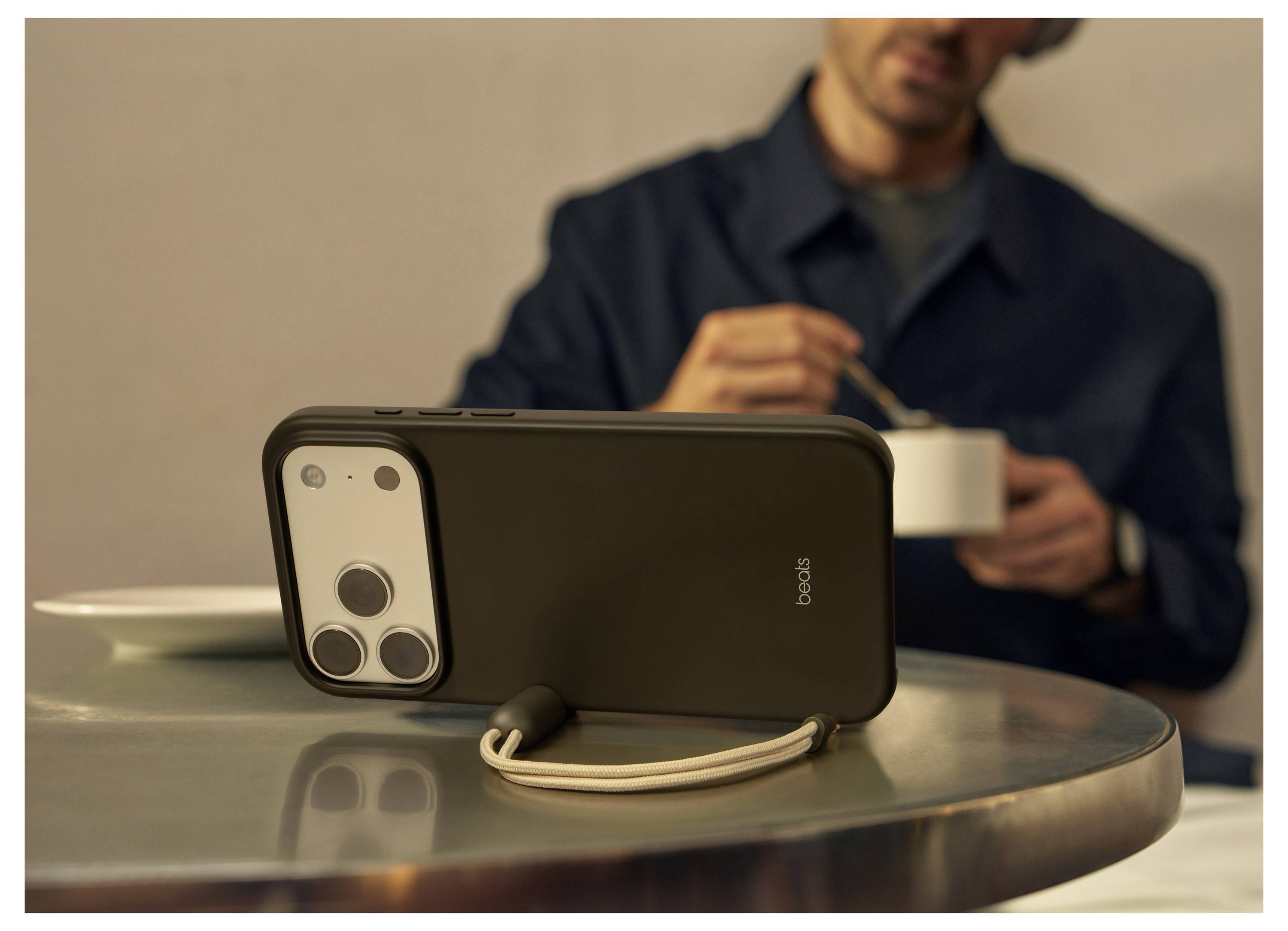 Ein Smartphone in einem Schutzgehäuse mit Handgelenkschlaufe liegt auf einem Tisch, während eine Person im Hintergrund eine Schüssel hält.