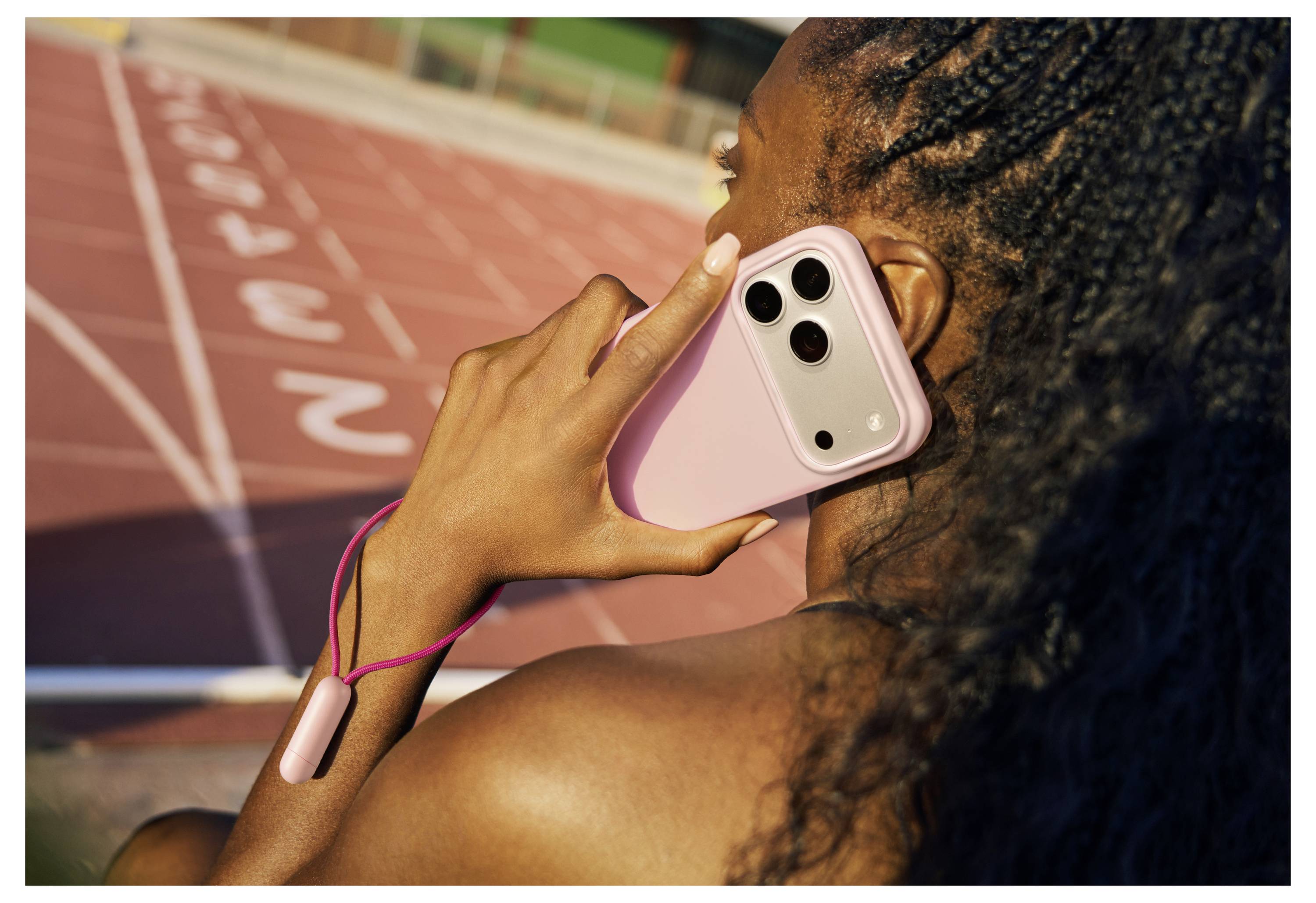 Eine Person hält ein rosa Smartphone an ihr Ohr, während sie neben einer Laufbahn steht und damit einen Fokus auf Fitness und Vernetzung andeutet.
