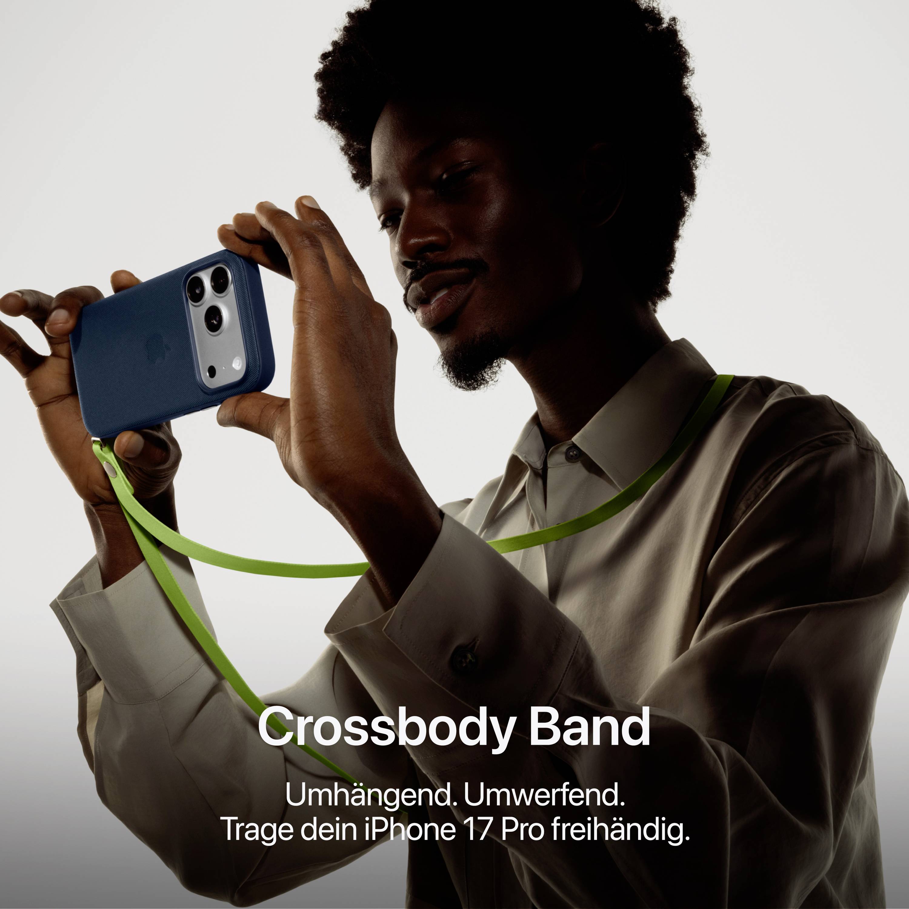 Ein Mann hält ein iPhone 17 Pro an einem grünen Crossbody-Band. Text: 'Crossbody Band. Umhängend. Umwerfend. Trage dein iPhone 17 Pro freihändig.'
