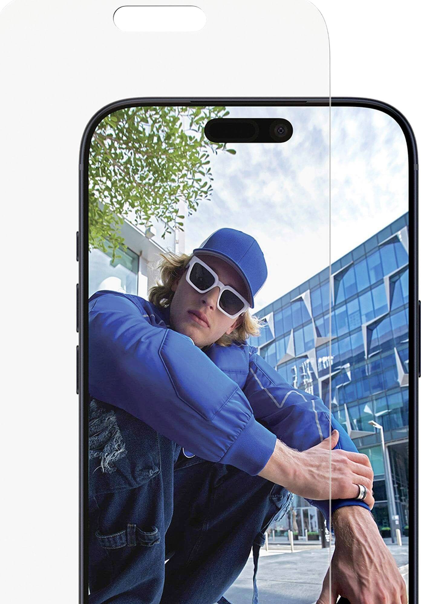 Person in blauer Jacke, Kappe und Sonnenbrille, die sich im Freien hockt, mit modernem Glasgebäude im Hintergrund, der auf einem Smartphone-Bildschirm reflektiert wird.