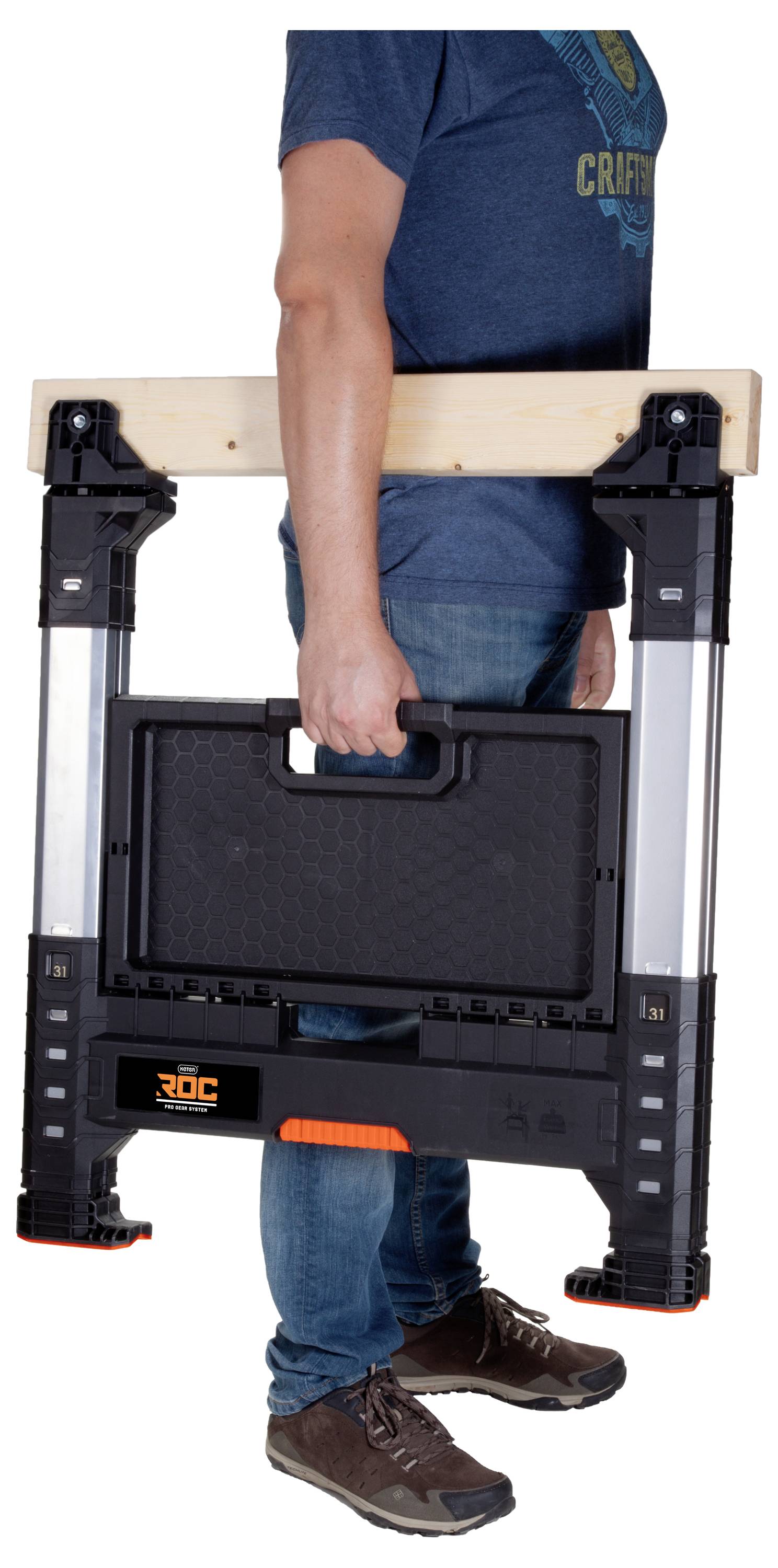 Eine Person, die eine zusammengefaltete mobile Werkbank am Griff trägt und dabei ihr kompaktes und leichtes Design für einfachen Transport demonstriert.