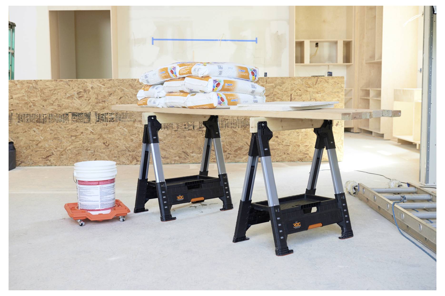 Ein Haufen Baumaterialien, darunter Säcke und ein Eimer, liegt auf einer Werkbank, die von Böcken gestützt wird, in einem teilweise fertiggestellten Raum.